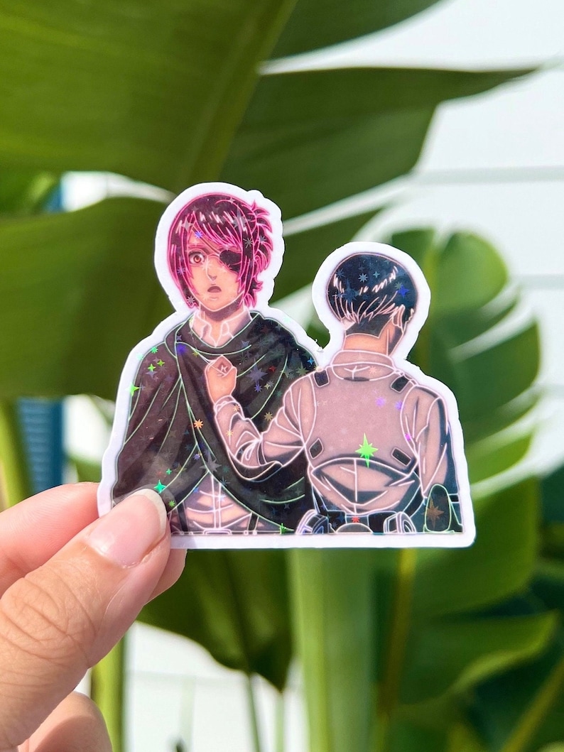 May include: A holographic sticker featuring two anime characters. The character on the left has pink hair and an eyepatch. The character on the right has black hair. Both characters are wearing military-style uniforms. The sticker has a glittery background.