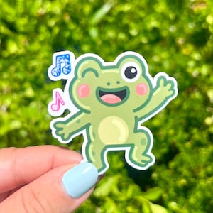 May include: A green frog sticker with a happy expression, winking and waving. The sticker has a white outline and features musical notes. The frog has a light yellow belly and pink cheeks. The background is a blurred green.