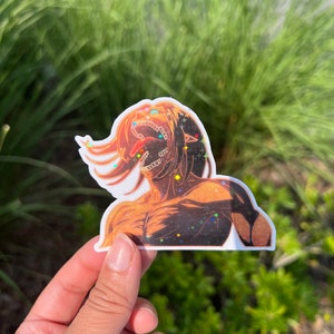 K&ouml;nnte beinhalten: Ein Aufkleber mit einem Charakter aus dem Anime Attack on Titan. Der Charakter ist ein Titan mit einem gro&szlig;en Maul und scharfen Z&auml;hnen. Der Aufkleber besteht aus klarem Vinyl mit holografischem Finish. Der Aufkleber ist mit Glitzer verziert.