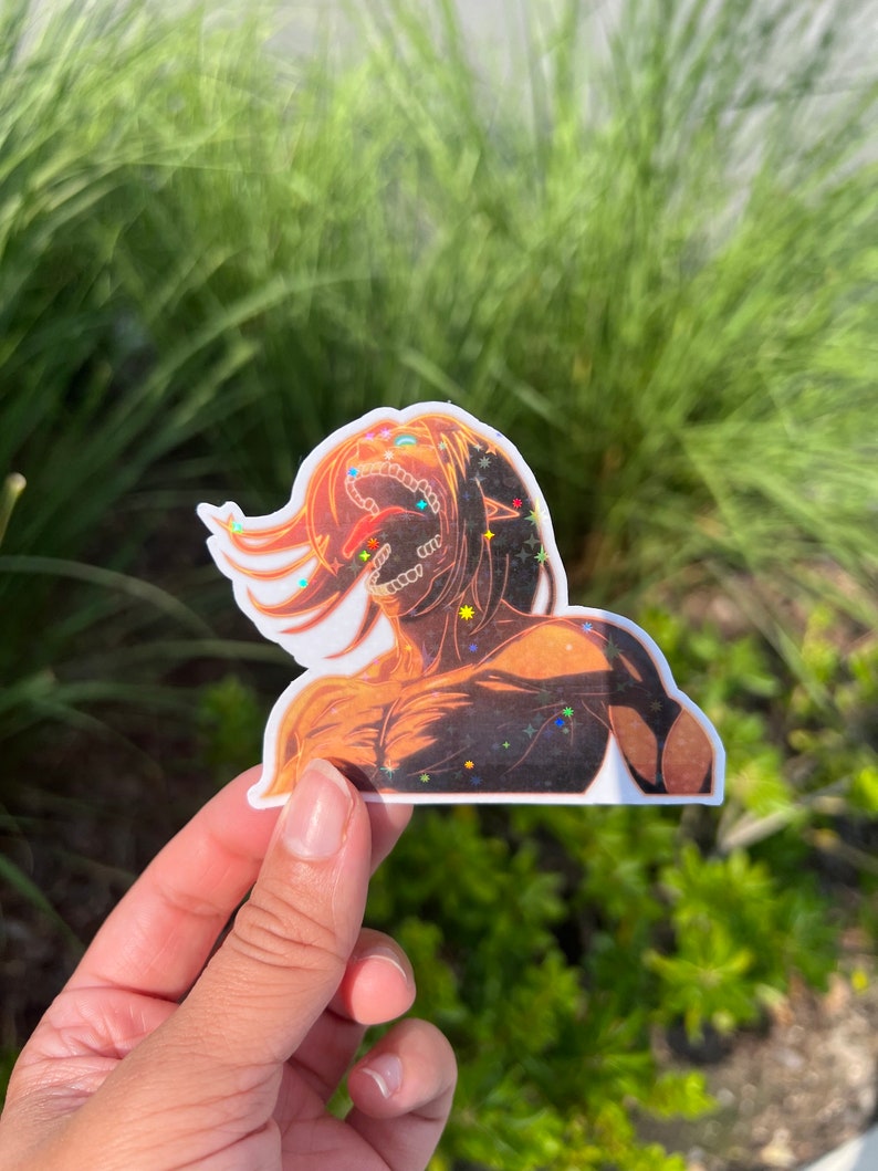 K&ouml;nnte beinhalten: Ein Aufkleber mit einem Charakter aus dem Anime Attack on Titan. Der Charakter ist ein Titan mit einem gro&szlig;en Maul und scharfen Z&auml;hnen. Der Aufkleber besteht aus klarem Vinyl mit holografischem Finish. Der Aufkleber ist mit Glitzer verziert.