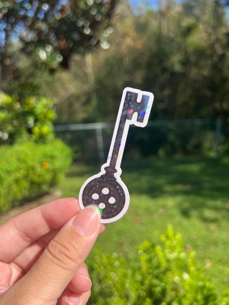 May include: A black and white iridescent key sticker with a round keyhole design. The keyhole has four small white circles.