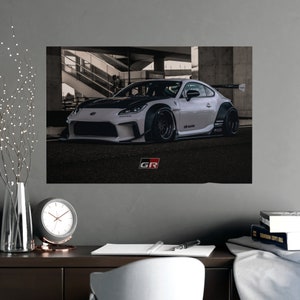 May include: A white and black modified Toyota GR 86 sports car with black wheels and a black and white GR logo on the front bumper. The car is parked in a concrete garage setting.