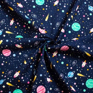 May include: Dark blue fabric featuring a space-themed design. The pattern includes planets, rockets, stars, and comets in pink, blue, yellow, and green. The fabric is likely cotton or a similar material, suitable for various craft projects.