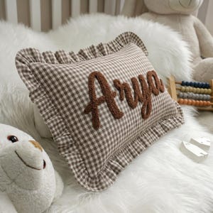 A brown and white checkered pillow with the name "Arya" embroidered on it, surrounded by a white fur-covered bed and a stuffed animal.