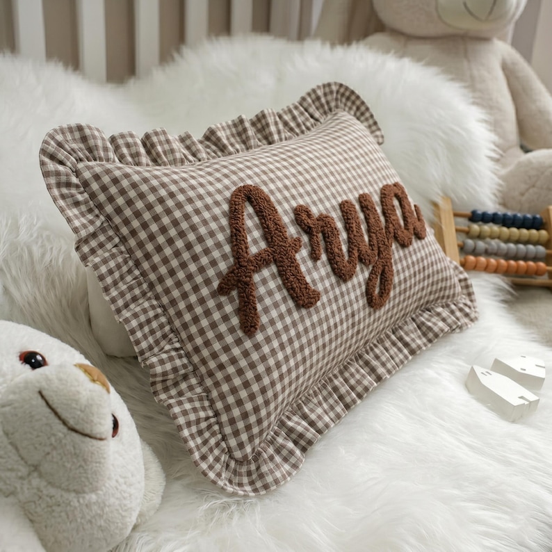 A brown and white checkered pillow with the name "Arya" embroidered on it, surrounded by a white fur-covered bed and a stuffed animal.