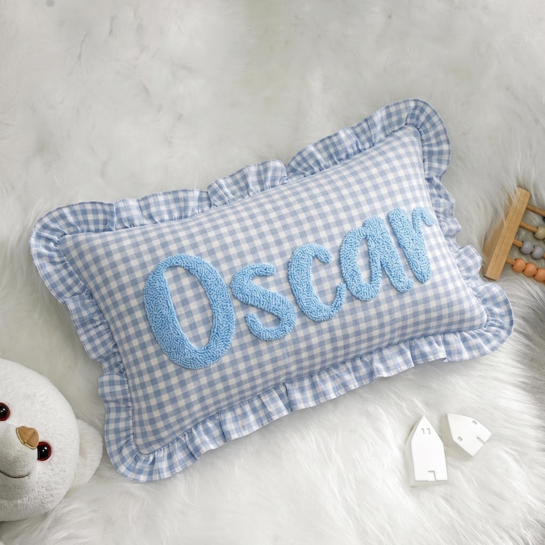 A blue and white gingham pillow with the name "Oscar" embroidered on it, placed on a white fur surface next to a stuffed animal and some small white objects.