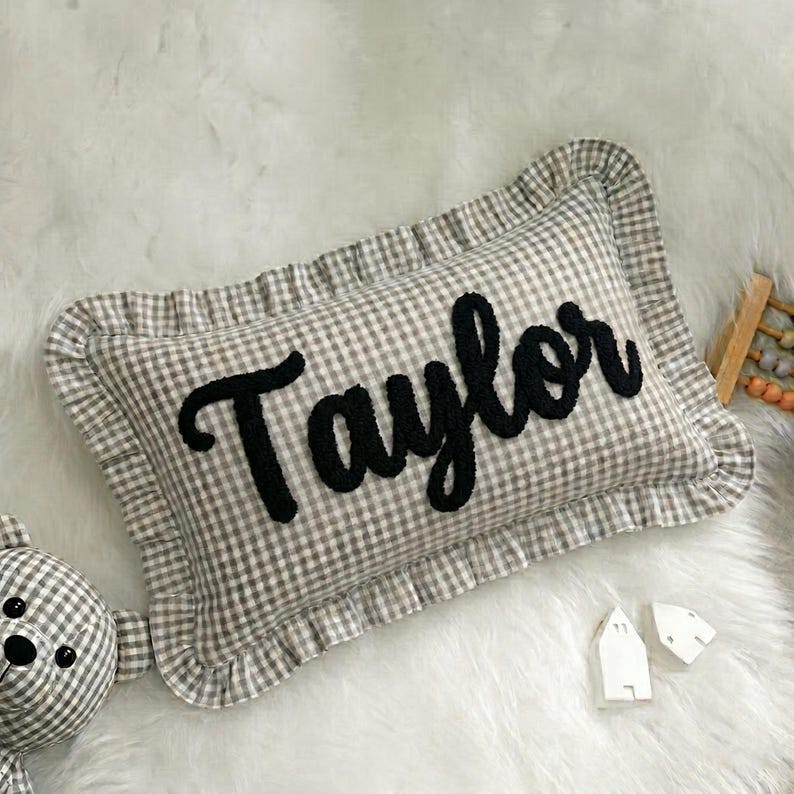 A decorative pillow with the name "Taylor" embroidered on it, placed on a white fur surface. There is also a teddy bear and some small white figurines in the background.