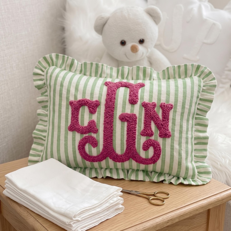 a decorative pillow with a monogrammed pink lettering on a green and white striped background, placed on a wooden surface. A white teddy bear is visible in the background, and there are some white cloth napkins and a pair of scissors on the surface.
