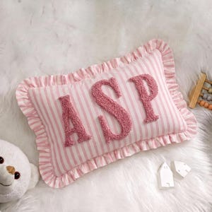 A pink and white striped pillow with the letters "ASP" on it, placed on a white fur surface. There is also a stuffed animal and some small white objects nearby.