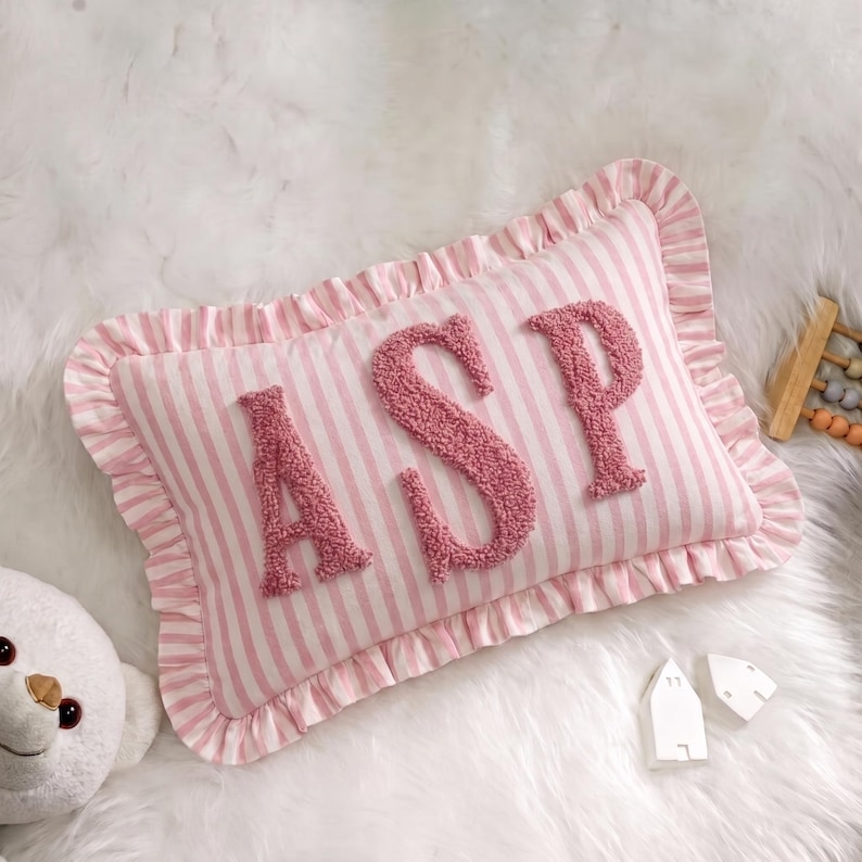 A pink and white striped pillow with the letters "ASP" on it, placed on a white fur surface. There is also a stuffed animal and some small white objects nearby.