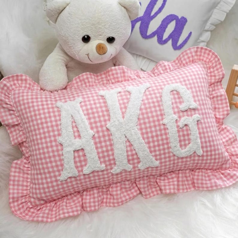 A white teddy bear sits next to a pink pillow with the initials "AKG" on it, placed on a white fur-covered surface.