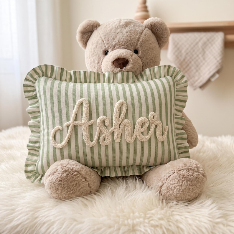 A stuffed teddy bear holding a green and white striped pillow with the name "Asha" written on it.