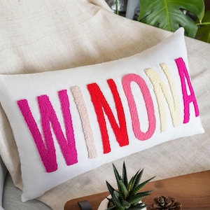 May include: White rectangular pillow with the name "WINONA" embroidered in vibrant colors. The letters are in shades of pink, red, and cream, creating a bold and eye-catching design. The pillow is placed on a neutral-toned surface.