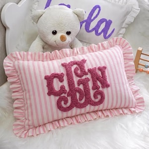 A white teddy bear sits on a pink and white striped pillow with the word "CJN" embroidered on it.
