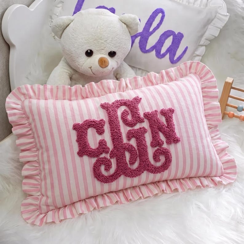 A white teddy bear sits on a pink and white striped pillow with the word "CJN" embroidered on it.