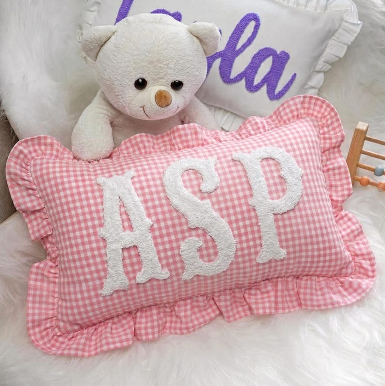 A white teddy bear sits on a pink and white checkered pillow with the letters "ASP" on it.