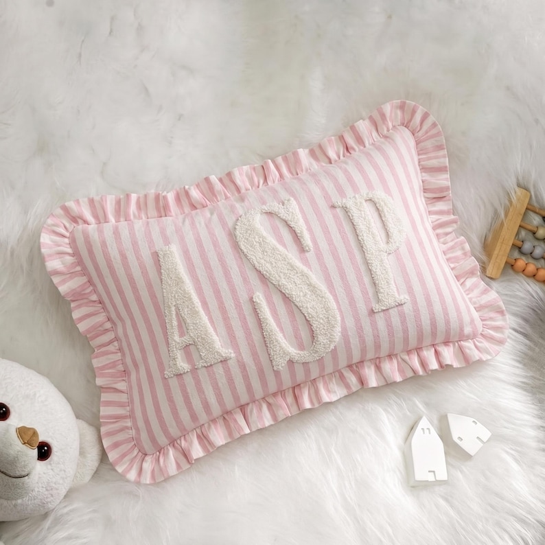 A pink and white striped pillow with the letters "ASP" on it, placed on a white fur surface. There is also a stuffed animal and some small white objects nearby.