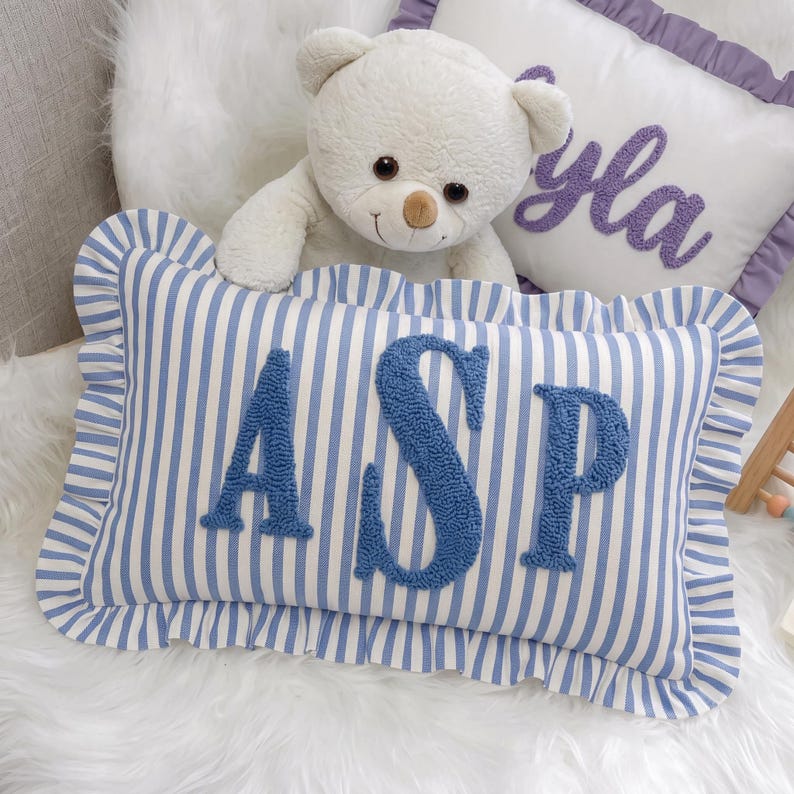 A white teddy bear sits on a white fur rug, with a blue and white striped pillow featuring the letters "ASP" in front of it.