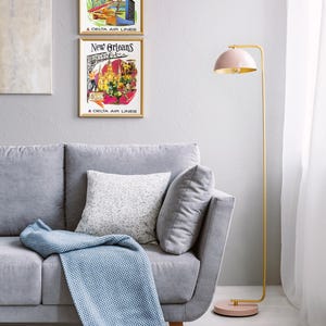 Puede incluir: Un sof&aacute; gris claro con un coj&iacute;n blanco moteado y una manta azul texturizada. Junto al sof&aacute; hay una l&aacute;mpara de pie con estructura de metal dorado, una pantalla en forma de c&uacute;pula rosa p&aacute;lido y una base circular a juego en rosa p&aacute;lido.