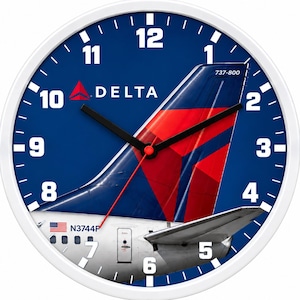 May include: A round wall clock with a white frame and a blue face featuring a Delta Airlines Boeing 737-800 tail fin graphic. The clock displays white numbers 1 through 12, black hour and minute hands, a red second hand, and the text Delta and 737-800.