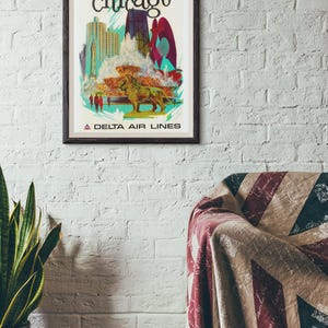 Puede incluir: Un p&oacute;ster de viaje enmarcado de Chicago con una ilustraci&oacute;n estilizada del horizonte de la ciudad, una gran fuente y una estatua de le&oacute;n de bronce. El texto dice Chicago en la parte superior y A Delta Air Lines en la parte inferior.
