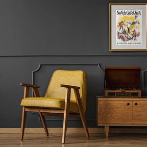 Puede incluir: Un p&oacute;ster de viaje vintage enmarcado con el texto North Carolina A Delta Air Lines, que representa figuras en un paisaje con flores. El p&oacute;ster se muestra en un marco dorado sobre una pared gris oscuro encima de un aparador de madera y un sill&oacute;n amarillo.