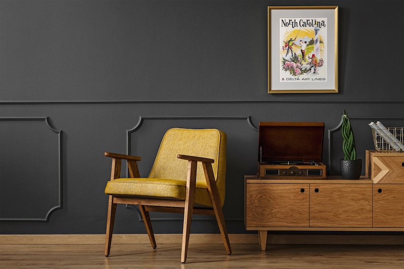 Puede incluir: Un p&oacute;ster de viaje vintage enmarcado con el texto North Carolina A Delta Air Lines, que representa figuras en un paisaje con flores. El p&oacute;ster se muestra en un marco dorado sobre una pared gris oscuro encima de un aparador de madera y un sill&oacute;n amarillo.