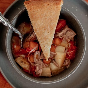 Puede incluir: Un tazón de estofado con carne desmenuzada, patatas, zanahorias y tomates cubierto con una rebanada de pan de maíz.