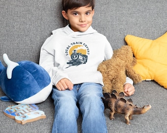 May include: A young person wearing a white hoodie with a vintage train graphic and the text "Local Train Expert". The person is sitting on a gray couch with a blue whale plush toy, a brown teddy bear, and a brown dinosaur plush toy.