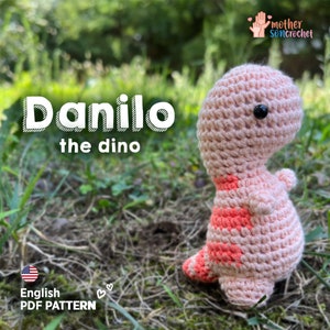 Puede incluir: Un dinosaurio de crochet rosa llamado Danilo el Dino, sentado en un lecho de hierba verde. El juguete tiene un ojo negro y una franja roja en la espalda. La imagen incluye el texto "Danilo el Dino" y "English PDF PATTERN".