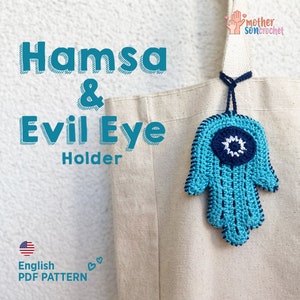 Op de afbeelding: Een gehaakte blauwe hamsa-handhanger met een wit en blauw boze oog in het midden. De hanger hangt aan een witte tote bag met de tekst "Hamsa & Evil Eye Holder" en "English PDF PATTERN" erop gedrukt.