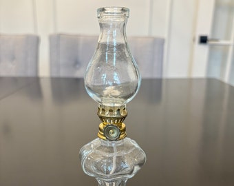 May include: A clear glass oil lamp with a brass-coloured metal base and a clear glass chimney. The lamp has a decorative, vintage design and is suitable for home decor. The lamp is approximately 15 cm tall.