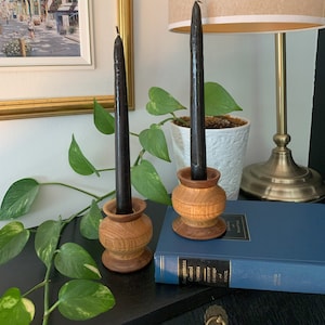 May include: Two black candles in wooden candle holders. The holders are made of light brown wood with a natural grain pattern. The candles are sitting on a black surface with a book and a plant in the background.