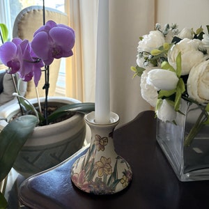 May include: A white candle sits in a ceramic candle holder with a floral design. The holder has a wide base and a narrow neck. The candle is tall and thin. There are flowers in the background.