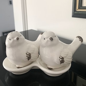 Pode incluir: Um conjunto de saleiro e pimenteiro de cerâmica branca em forma de pássaros. Os pássaros estão empoleirados em uma bandeja de cerâmica branca. Os pássaros têm detalhes marrons em suas asas e caudas.