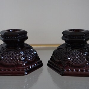 Pode incluir: Dois castiçais de vidro vermelho escuro com desenhos intrincados. Os castiçais são hexagonais e têm uma borda de contas.