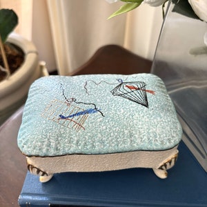May include: A small, square, blue and white ceramic trinket box with gold trim and three feet. The box has a design of a kite and other abstract shapes.