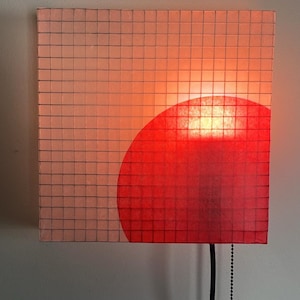 May include: Square wall lamp with a grid pattern and a red semi-circle design. The lamp is lit, emitting a warm glow. A black cord and pull chain hang from the bottom.