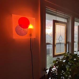 Könnte beinhalten: Eine Wandlampe mit einem roten Kreis und einem hellblauen Kreis auf einem gitterartigen Hintergrund. Die Lampe ist beleuchtet und wirft einen orangefarbenen Schein. Ein Fenster mit einer Glasmalerei hängt vor einem Sonnenuntergang.