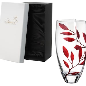 May include: A clear glass vase with a red leaf design. The vase is in a white box with a black lid. The box has the text "Anna's Exclusive Designs" on it.