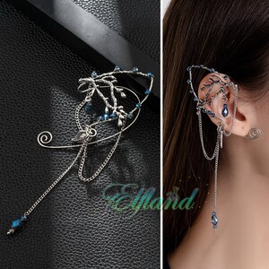 Könnte beinhalten: Ein silberfarbener Ohrring mit einer astähnlichen Struktur, verziert mit kleinen blauen Perlen und einer hängenden Kette mit einer tropfenförmigen blauen Perle. Das Metalldesign enthält einen Spiralakzent und der Text Elfland ist zu sehen.