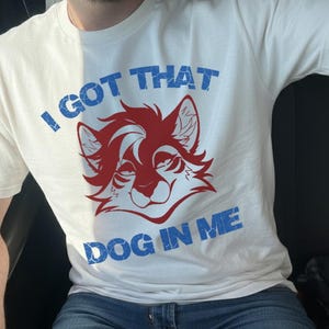 Puede incluir: Camiseta blanca con el texto "I GOT THAT DOG IN ME" en azul y una cara de perro de dibujos animados roja y blanca. La camiseta está hecha de un material suave.