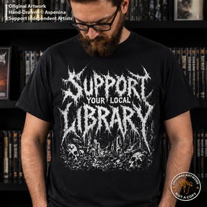 May include: A black short-sleeved t-shirt featuring a white graphic print with the text "SUPPORT YOUR LOCAL LIBRARY" in a jagged, distressed font. The design includes detailed illustrations of skulls and ruins at the bottom of the graphic.