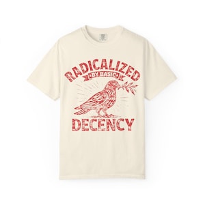 以下が含まれることがあります： クリーム色のTシャツ。赤色のグラフィックは、枝を加えた鳩のデザインで、「RADICALIZED BY BASIC」の文字が上に、「DECENCY」の文字が下に配置されています。Tシャツは半袖で、クルーネックです。