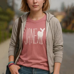 May include: A dusty rose t-shirt featuring the word "JANE" in white, stylized text, and a white deer graphic. The shirt is worn by a person wearing a grey zip-up hoodie and blue jeans.