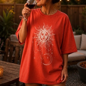 May include: A bright orange short-sleeved t-shirt featuring a large white graphic print of a sun with a human face, surrounded by radiating rays, dangling star charms, and a crescent moon positioned below the sun on the center of the chest.