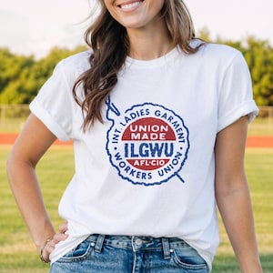 Puede incluir: Camiseta blanca con un diseño gráfico circular en azul y rojo que dice "INT. LADIES GARMENT WORKERS UNION". La camiseta tiene las mangas dobladas y está hecha de un material suave. El diseño incluye las letras "ILGWU" y "AFL-CIO".