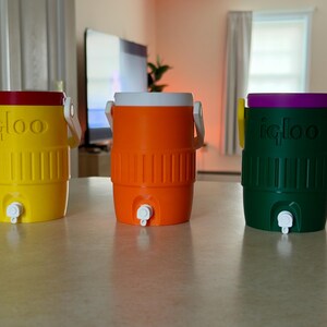 May include: Three colorful Igloo water coolers. The coolers are yellow with a red lid, orange with a white lid, and green with a purple lid. Each cooler has a white handle and a spigot for dispensing liquids.