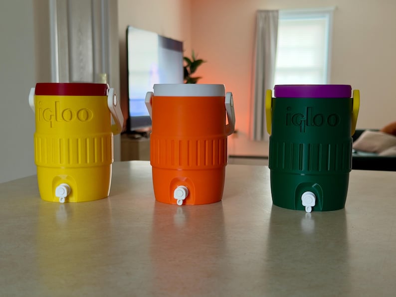 May include: Three colorful Igloo water coolers. The coolers are yellow with a red lid, orange with a white lid, and green with a purple lid. Each cooler has a white handle and a spigot for dispensing liquids.