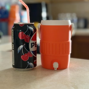 May include: A black cherry-flavored Pepsi can with a pink straw next to an orange and white cooler. The can features a cherry design and the Pepsi logo. The cooler has a spigot and a white lid.